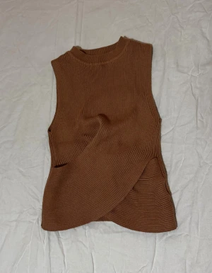 Brun ribbad omlott linnetopp - Säljer en brun ärmlös linnetopp med ribbad struktur och snygg omlott-design framtill. Toppen har en hög hals och är gjord i ett stickat material som känns mjukt och skönt mot huden. Perfekt för dig som gillar stilrena och trendiga plagg.