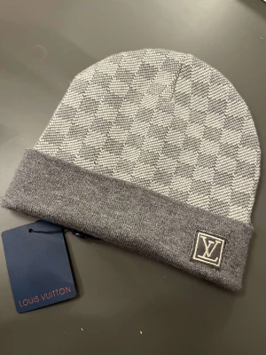 Grå rutig mössa - Snygg grå mössa med rutigt mönster och uppvikt kant. Stickad i mjukt material som håller dig varm och stilren under kalla dagar. Perfekt accessoar för dig som gillar designer och streetstyle.