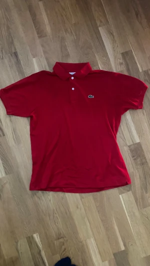 Röd pikétröja från Lacoste - Snygg röd pikétröja från Lacoste med klassisk krage och två knappar framtill. Ikoniska gröna krokodilloggan broderad på bröstet. Tröjan är tillverkad i mjuk bomull och har korta ärmar, perfekt för en chill och sportig look. Storlek 4, passar ungefär M herr! Vid fler frågor/funderingar tveka inte på att skriva!