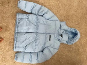 Ljusblå pufferjacka från Hoodrich - Säljer en ljusblå pufferjacka från Hoodrich med stor logga på ryggen och mindre tryck på bröstet. Jackan har huva, dragkedja framtill och två snedställda fickor. Perfekt för kalla dagar när du vill ha en clean och modern look.