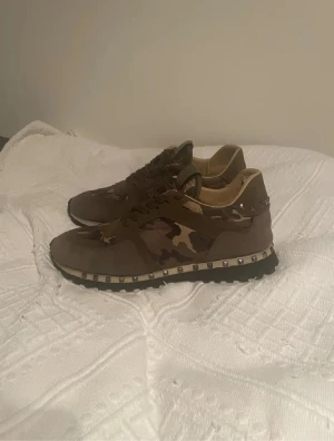 Valentino Rockrunner  - Säljer ett par Valentino Rockrunner sneakers i grön och beige med camouflage-mönster. Skorna har snygga nitar runt sulan, snörning och en platt sula. Materialmix av mocka och skinn ger en lyxig känsla. Perfekt för dig som vill sticka ut med coola detaljer och streetstyle.