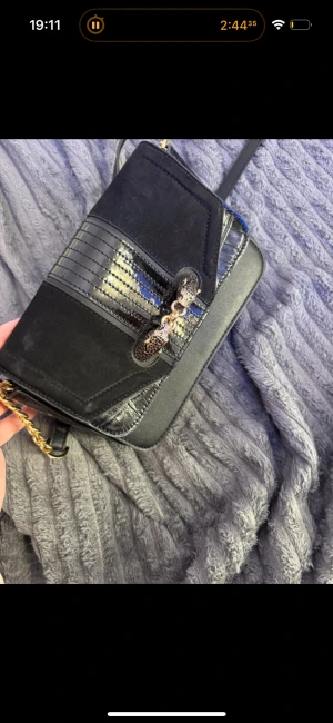 Svart axelväska med guldkedja - Snygg svart axelväska med guldfärgad kedja och detaljer. Väskan har en mix av mocka och skinn, samt ett coolt mönster på locket. Insidan är rymlig med flera fack, perfekt för mobilen och småsaker. Stilren och trendig design som lyfter din outfit.