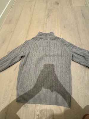 Grå polotröja från Polo Ralph Lauren - Snygg grå polotröja från Polo Ralph Lauren med kabelstickat mönster och ribbad krage. Tröjan har långärmade ärmar och en half zip-dragkedja framtill. Perfekt för dig som gillar klassisk stil med en modern touch. Aldrig andvänt den beställde den och så var den för liten från kidsbranstore ny pris 1795