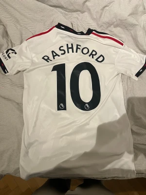 Manchester United Rashford #10 22/23 bortaställ  - Säljer en officiell Manchester United bortatröja från Adidas med Rashford och nummer 10 på ryggen. Tröjan är vit med svarta och röda detaljer, klubbmärke på bröstet och TeamViewer-logga. Tillverkad i lätt polyester med Aeroready-teknologi för extra komfort.