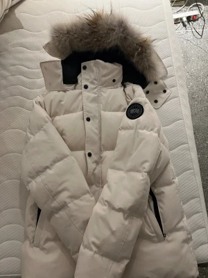 Vit Wyndham från Canada Goose - Snygg vit dunjacka från Canada Goose med svart logotyp på ärmen och stor huva med fluffig päls. Jackan har svarta knappar, dragkedja och praktiska fickor. Perfekt för kalla vinterdagar och riktigt varm med sin tjocka vaddering.