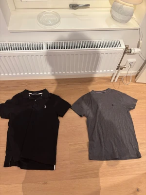 Svart och grå t-shirt från U.S. Polo Assn. - Två snygga t-shirts från U.S. Polo Assn. Den ena är svart med krage och knappar, klassisk pikéstil. Den andra är grå med rund hals och diskret logga på bröstet. Båda är i mjuk bomull och har korta ärmar, perfekta för en chill och stilren look.