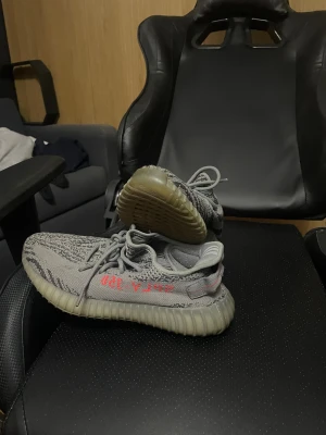Adidas Yeezy Boost 350 V2 Beluga - Säljer ett par Adidas Yeezy Boost 350 V2 Beluga med grå och mörkgrå stickad ovandel, orange 'SPLY-350' text på sidan och en tjock, ribbad platt sula i beige. Skorna har snörning och en rund tå, tillverkade i syntet och stickad textil för en skön och modern look.