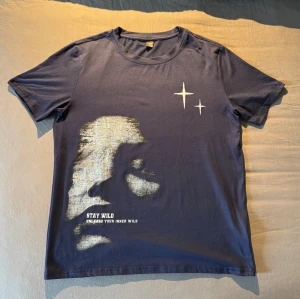 Mörkblå t-shirt med tryck och text - Mörkblå t-shirt med coolt tryck av ett ansikte i silver och texten 'STAY WILD UNLEASH YOUR INNER WILD' på framsidan. Två vita stjärnor tryckta på bröstet. Klassisk passform med rund hals och korta ärmar. Perfekt för dig som vill sticka ut lite extra. Storlek S. Använd cirka en gång. Det är bara att höra av sig om man har någon fråga :)