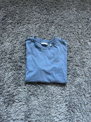 Blå t-shirt från GANT med broderad logga - Snygg blå t-shirt från GANT med rund hals och liten broderad logga på bröstet. Tillverkad i mjuk bomull som känns skön mot huden. Perfekt för dig som gillar klassisk stil med en twist.