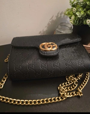 Svart axelväska i skinn från Gucci - Snygg svart axelväska från Gucci i präglat skinn med GG-logga och guldfärgad kedja. Klassisk design med lock och magnetlås, perfekt storlek för det viktigaste. Väskan har exklusiva detaljer och känns riktigt lyxig.