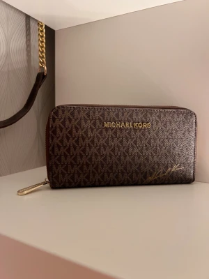 Brun Mikael kors plånbok  - Snygg brun plånbok från Michael Kors med klassiskt MK-logomönster över hela ytan. Plånboken har guldfärgad dragkedja och flera fack för kort och mynt. Materialet är skinn och designen är stilren med guldfärgade detaljer. Aldrig använt