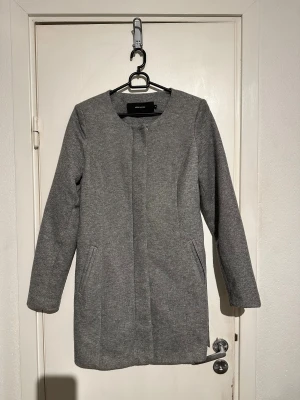 Grå kappa från Vero Moda med bälte - Stilren grå kappa från Vero Moda med rak passform och avtagbart bälte i midjan. Jackan har dold knäppning framtill, två fickor och är helfodrad. Perfekt för dig som gillar en clean look och vill ha något som funkar till många outfits.