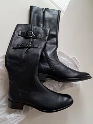 Svarta stövlar från Gabor i skinn - Snygga svarta stövlar från Gabor med rund tå och låg klack. Tillverkade i mjukt skinn med dragkedja på insidan och två coola spänndetaljer upptill. Perfekt till höst och vinter för en stilren look.