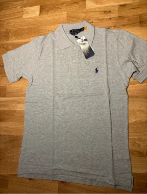  pikétröja Polo Ralph Lauren - Snygg grå pikétröja från Polo Ralph Lauren med klassisk krage och två knappar framtill. Tröjan har korta ärmar och den ikoniska broderade loggan i mörkblått på bröstet. Perfekt för en clean och stilren look.