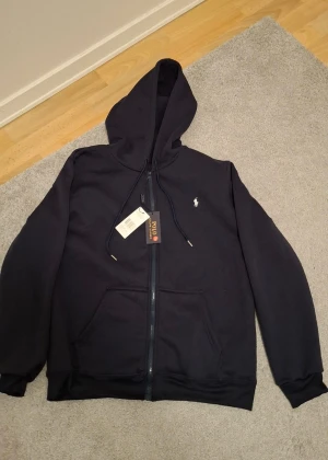 Svart hoodie från Polo Ralph Lauren - Marinblå hoodie från Polo Ralph Lauren med dragkedja och huva. Klassisk design med logotyp på bröstet, snörning i huvan och två fickor framtill. Perfekt för chill dagar och enkel att matcha med jeans eller joggers.