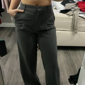 Mörkgrå vida kostymbyxor - Snygga mörkgrå kostymbyxor med hög midja och vida ben. Byxorna har pressveck framtill och stängs med knapp och dragkedja. Perfekta för en stilren och trendig look. byxorna är helt nya från bik bok!! Köpta för 599kr säljer för 199