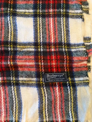 Rutig halsduk från Burberrys i ull - Snygg rutig halsduk från Burberrys i 100% lammull, tillverkad i England. Färgerna är vit, röd, blå, gul och svart med klassiskt rutmönster och fransar i ändarna. Perfekt för att ge din outfit en cool brittisk touch.
