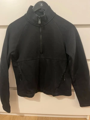 Svart half zip tröja Sail Racing M - Svart långärmad tröja från Sail Racing i storlek M. Tröjan har hög krage, half zip-dragkedja framtill och dragkedjeförsedda fickor på sidorna. Tillverkad i mjuk bomullsmix, perfekt för kyligare dagar. Snygg och enkel design med Sail Racing-logga bak. I storlek M för barn