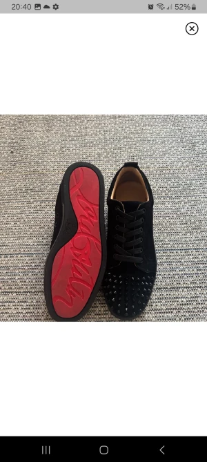 Svarta Louboutin sneakers med nitar - Säljer ett par svarta sneakers från Christian Louboutin i mocka med coola svarta nitar på tån och den ikoniska röda sulan. Skorna har snörning och en rund tå, vilket ger en edgy och lyxig vibe. Perfekta för dig som vill sticka ut med din stil.