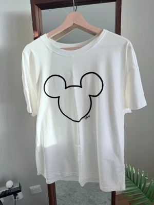 Vit H&M t-shirt med Mickey Mouse-tryck - Vit t-shirt från H&M Disney med en stor, svart kontur av Mickey Mouse-huvudet på bröstet. Klassisk rund hals och korta ärmar. Tillverkad i mjuk bomull för en skön och avslappnad känsla. 