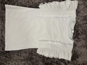 Vit blus med volang från Amisu - Supersöt vit blus från Amisu i storlek S. Toppen har breda axelpartier med smock och volangdetaljer som ger en trendig och feminin vibe. Materialet är mjukt och ribbat, perfekt för dig som gillar stilrena och romantiska plagg. Rund halsringning och ärmlös design.