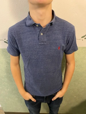 Blå pikétröja Polo Ralph Lauren M - Snygg blå pikétröja från Polo Ralph Lauren i slim fit-modell. Tröjan har korta ärmar, klassisk krage och knappar framtill. Ikoniska röda loggan broderad på bröstet. Perfekt för en clean och stilren look.