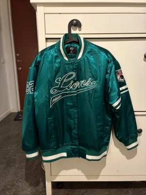 Grön bomberjacka Lions Athletic - Snygg grön bomberjacka från new yorker med broderad Lions Athletic-text på bröstet och coola patches på ärmarna. Jackan har vita detaljer vid muddar och krage, samt tryckknappar framtill. Materialet är glansigt polyester och modellen är loose fit.