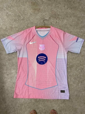 Barça x Nike rosa fotbollströja - Säljer en FC Barcelona x Nike fotbollströja i ljusrosa och lila toner med korta ärmar. Tröjan har Spotify-logga på bröstet, UNHCR-tryck på ryggen och klubbmärke framtill. Materialet är lätt och andas, perfekt för match eller träning.