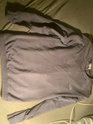 Grå långärmad tröja från Lacoste - Snygg grå långärmad tröja från Lacoste med klassisk rund hals och den ikoniska gröna krokodilen broderad på bröstet. Tröjan är tillverkad i mjuk bomull och har ribbade muddar vid ärmslut och nederkant. Perfekt för dig som gillar stilrena och enkla plagg.