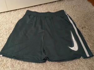 Nike Dri-Fit träningsshorts mörkgrön - Säljer ett par mörkgrå Nike Dri-Fit träningsshorts med vit rand längs sidan och stor vit Swoosh-logga. Shortsen har elastisk midja och är tillverkade i ett lätt, snabbtorkande syntetmaterial som passar perfekt för sport och träning.