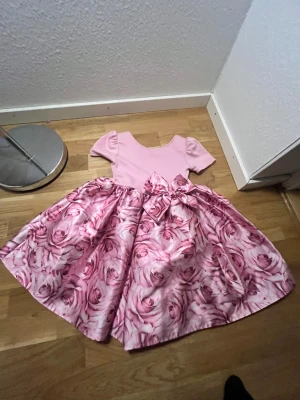 Rosa blommig klänning med rosett - Supersöt rosa klänning med kort ärm och vidd i kjolen. Överdelen är enfärgad ljusrosa och kjolen har ett stort blommönster i olika rosa nyanser. Stor rosett i midjan framtill ger extra wow-känsla. Perfekt för dig som älskar romantiska vibes och blommiga prints.