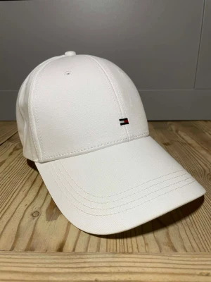 Vit keps från Tommy Hilfiger - Snygg vit keps från Tommy Hilfiger med klassisk böjd skärm och diskret broderad logga i rött, vitt och blått framtill. Kepsen är tillverkad i bomull och har ventilerande hål upptill. Perfekt för dig som gillar stilrena accessoarer.