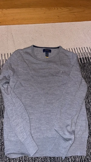 Grå stickad tröja från Polo Ralph Lauren - Snygg grå stickad tröja från Polo Ralph Lauren med rund halsringning och diskret broderad logga på bröstet. Tröjan har långa ärmar och ribbade muddar för en klassisk look. Perfekt till jeans eller chinos.