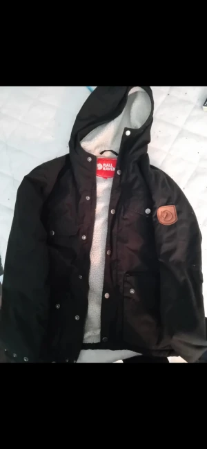 Fjällräven Greenland Winter Jacket - Fjällräven Greenland Winter Jacket   Svart jacka från Fjällräven med teddyfoder och huva. Jackan har flera fickor med tryckknappar, brunt Fjällräven-märke på ärmen och svensk flagg vid nederkanten. Yttermaterialet är slitstark polyester och bomull, perfekt för kalla dagar. Väldigt bra skick och inga skavanker vad man ser. Kostar 5000kr i nypris. Storlek är XS. Använd ca 2 månader förra vintern.