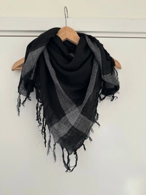 Svart och grå rutig halsduk med fransar - Snygg svart halsduk med grått rutmönster och fransar längs kanterna. Den är stor och fyrkantig, perfekt att svepa runt halsen eller använda som sjal. Materialet är tunt och vävt, vilket gör den lätt och luftig. Passar dig som gillar en cool och enkel stil.