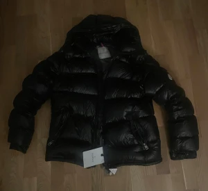 Moncler maya  - Snygg svart dunjacka från Moncler med glansig finish, huva och flera dragkedjeförsedda fickor. Jackan har klassisk Moncler-logga på ärmen och coolt serietryck på insidan. Perfekt för kalla dagar när du vill hålla stilen. Skicken är bra. Pris kan diskuteras.