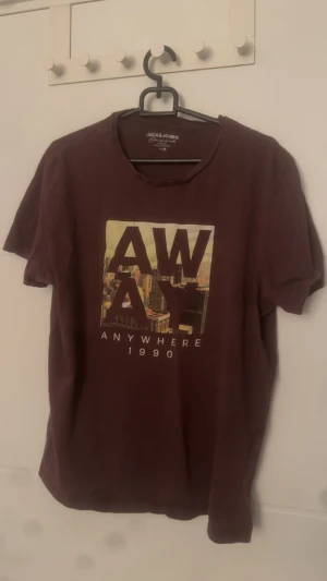 Vinröd t-shirt Jack & Jones AWAY - Vinröd t-shirt från Jack & Jones med tryck framtill där det står 'AWAY ANYWHERE 1990' och en stadssilhuett i bakgrunden. Klassisk rund hals och korta ärmar. Perfekt för dig som gillar streetstyle och snygga prints.