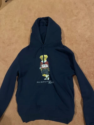 Marinblå hoodie Polo Ralph Lauren - Marinblå hoodie från Polo Ralph Lauren med ikoniska Polo Bear-trycket på bröstet. Tröjan har huva med snörning och långa ärmar. Mjuk insida och klassisk passform, perfekt för dig som gillar streetstyle och vill ha något bekvämt och snyggt.