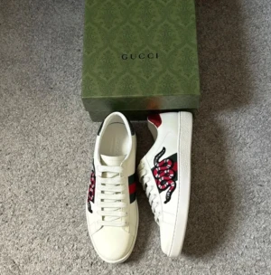 Gucci Ace sneakers med orm brodyr - Gucci Ace sneakers i vitt skinn med ikonisk röd och svart orm brodyr på sidan, gröna och röda ränder samt metallic-detaljer i rött och grönt på hälen. Klassisk låg modell med snörning och rund tå. Perfekta för dig som vill sticka ut med lyxig streetstyle.
