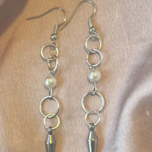 Långa silverörhängen med pärldetalj - Säljer ett par långa örhängen i silverfärg med flera ringar, vita pärlor och spetsiga metallhängen längst ner. Perfekta för dig som gillar statement-smycken med en cool vibe.