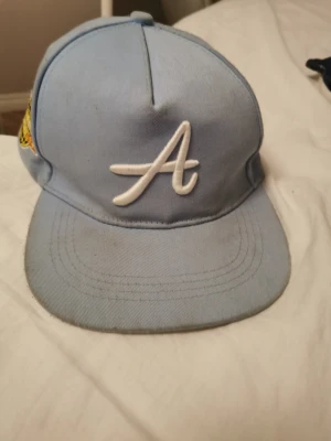 Ljusblå keps med broderi Los Angeles - Snygg ljusblå keps med vit broderad A på fronten och ett coolt broderat märke på sidan med texten 'Los Angeles Airport 1939' i gult, svart och rött. Klassisk form med böjd skärm, perfekt för att lyfta din streetstyle.