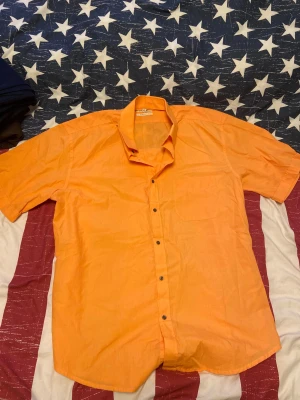 Orange kortärmad skjorta Summer Style - Fräsch orange skjorta från Summer Style med korta ärmar och klassisk krage. Skjortan har svarta knappar framtill och en bröstficka. Perfekt för varma dagar och har en clean, enkel look som sticker ut. Materialet känns lätt och luftigt. Skjortan är det inget fel på, den är använd i gott skick. Storlek 45/46 alltså motsvarar storlek L eller XL. 