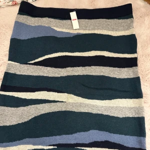 Stickad mönstrad kjol XL - Säljer en stickad kjol i storlek XL med coolt vågigt mönster i mörkgrön, grå, blå och svart. Kjolen har en rak form och är mjuk och skön. Obs har en lite fläck på framsidan som kom med när den köptes aldrig använd