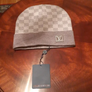 Rutig beige mössa från Louis Vuitton - Snygg beige mössa från Louis Vuitton med klassiskt rutigt mönster och LV-logga på kanten. Mössan har en bred ribbad kant och är tillverkad i mjukt material som känns lyxigt. Perfekt för dig som vill ha en exklusiv och stilren look.