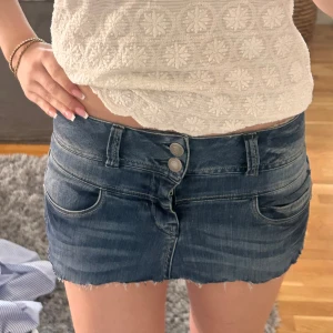 Jeanskjol mini från Pulz - Jeanskjol från Pulz med rå kant nertill och klassiska fem fickor. Kjolen har dubbla knappar framtill, bälteshällor och slitna detaljer för en cool look. Perfekt för dig som gillar denim och vill ha en edgy stil.