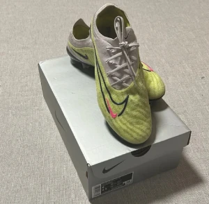 Nike Phantom GX fotbollsskor - Nike Phantom GX fotbollsskor i neongul och vit med svarta och rosa detaljer. Skorna har snörning, rund tå och platt sula med dobbar för gräsplan. Ovandelen är tillverkad i syntetmaterial med mönstrad struktur för bättre bollkontroll. Säljes med originalkartong och Nike phantom gympapåse. Tveka inte att ställa frågor eller bud.