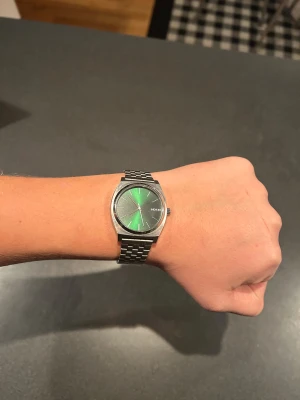 Grön Nixon klocka med metallarmband - Snygg klocka från Nixon med grön urtavla som har en cool solreflex-effekt. Klockan har ett klassiskt runt silverfärgat boett och ett stilrent metallarmband i silver. Minimalistisk design med tydliga index och diskret Nixon-logga på urtavlan. Perfekt för dig som gillar stilrena accessoarer.