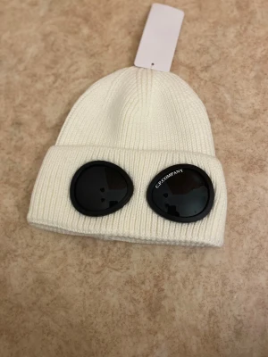 Vit C.P. Company goggle-mössa - Vit ribbstickad mössa från C.P. Company med ikoniska svarta goggle-linser framtill. Klassisk uppvikt kant och cool streetwear-vibe. Tillverkad i mjukt stickat material som håller dig varm och sticker ut i mängden.