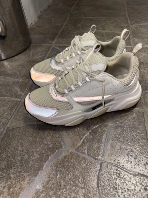 Dior B22 sneakers i vit och silver - Säljer ett par Dior B22 sneakers i vit och silver med chunky sula och meshdetaljer. Skorna har reflekterande paneler, snörning och en modern, sportig vibe. Märket syns på sidan och innersulan. Perfekt för dig som gillar exklusiva sneakers med statement-look.