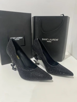 Saint Laurent svarta pumps med strass - Säljer ett par ikoniska Saint Laurent pumps i svart med glittrande strass över hela skon och spetsig tå. Klacken är formad som YSL-loggan vilket ger en riktigt lyxig vibe. Ovandelen är i skinn och insidan har guldfärgad Saint Laurent-text. Perfekta för dig som vill sticka ut.